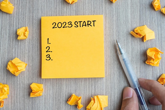 2023 start word on yellow note with businessman holding pen and crumbled paper on wooden table background new year resolutions strategy and goal concept