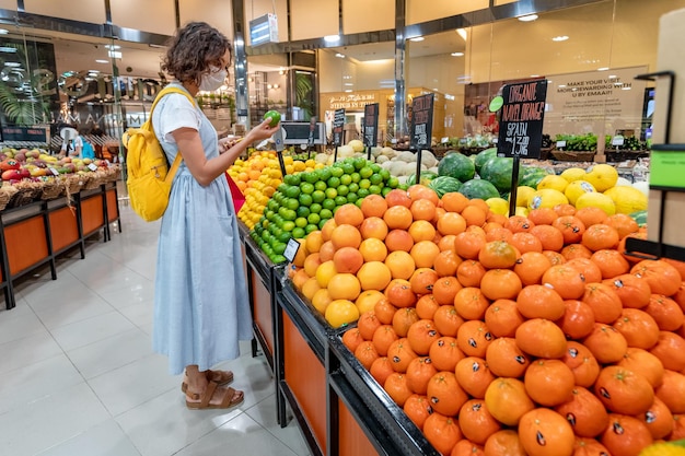 2021年2月23日 - スーパーマーケットの食料品店でマスクをかぶった女性顧客がショッピングをしライムとレモンの果物を摘み取ります