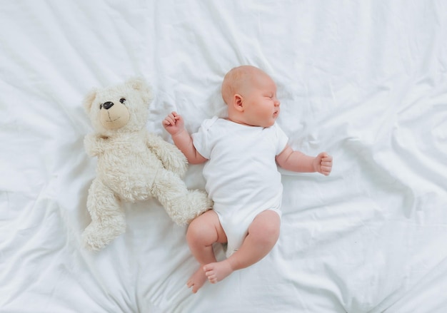 A 2monthold baby in a white bodysuit is lying at home on a white bed next to a teddy bear view from above newborn