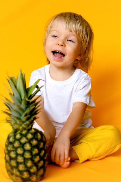 Photo a 2yearold child with blond hair in a white tshirt and yellow pants with a ripe big pineapple on