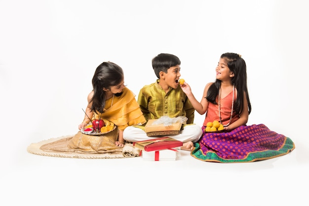 3 indian kids or siblings in traditional wear sitting on sofa or white background, holding gifts and sweets or laddu