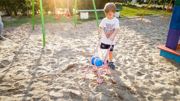 3 years old toddler boy playing with pram for dolls on the playground