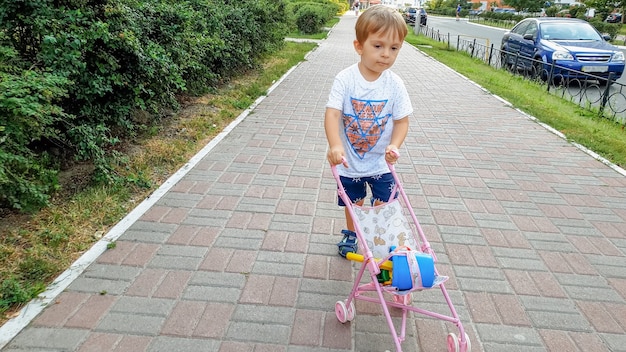 Ragazzo del bambino di 3 anni che cammina con la carrozzina del giocattolo sulla via. ragazzo che gioca con i giocattoli per le ragazze.