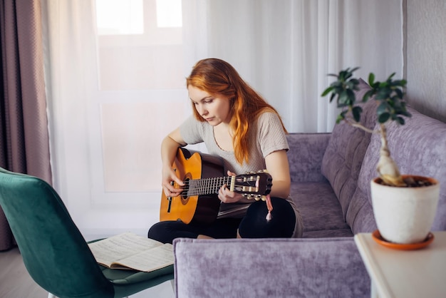 30 женщина играет на гитаре, сидя на диване в домашнем интерьере. Прекрасная молодая девушка учится играть на гитаре с нотами. Образ жизни, хобби, досуг, концепция развития талантов.