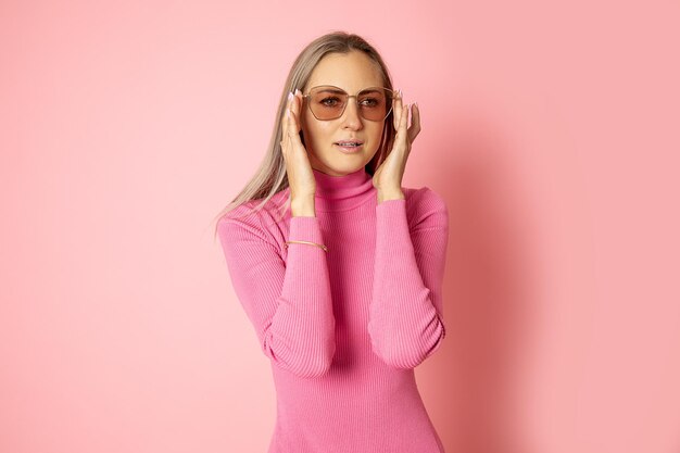 3035 years slender woman in bright pink dress touching sunglasses with hands on pink background