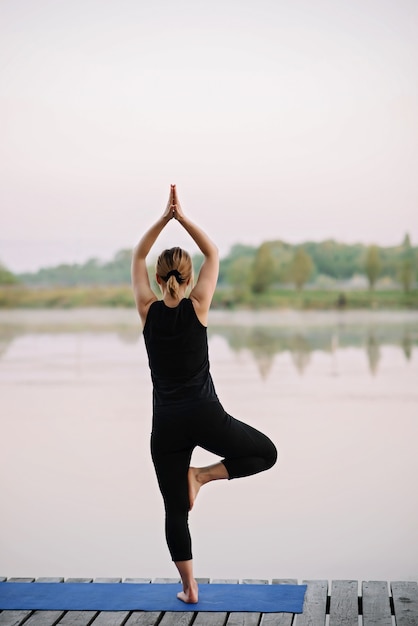 A 36-year-old young Caucasian woman practices yoga outdoors near a river on a wooden pier in the morning