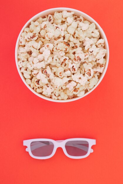 3d glasses and a paper cup with popcorn are isolated on a red background, a view from above