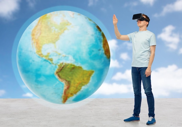 3d technology, virtual reality, entertainment, cyberspace and people concept - happy man with virtual reality headset or 3d glasses playing game and touching earth globe over sky and clouds background