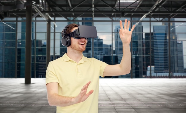 3d technology, virtual reality, entertainment and people concept - happy young man with virtual reality headset or 3d glasses playing game over industrial empty room and city panorama background