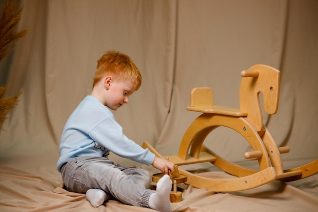 3 歳の赤毛の男の子が木の馬をブランコにして遊んでいて、素敵な服を着ています。