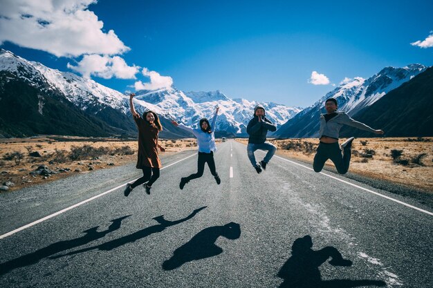 写真 4人の友達が雪に覆われた山の後ろの道路で高くジャンプする 新しい熱心なロードトリップ