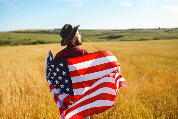 4 juli. Vier juli. Amerikaan met de nationale vlag. Amerikaanse vlag. Onafhankelijkheidsdag. Patriottische vakantie. De man draagt een hoed, een rugzak, een overhemd en een spijkerbroek.