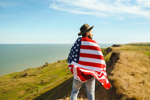 4 juli. Vier juli. Amerikaan met de nationale vlag. Amerikaanse vlag. Onafhankelijkheidsdag. Patriottische vakantie. De man draagt een hoed, een rugzak, een overhemd en een spijkerbroek.