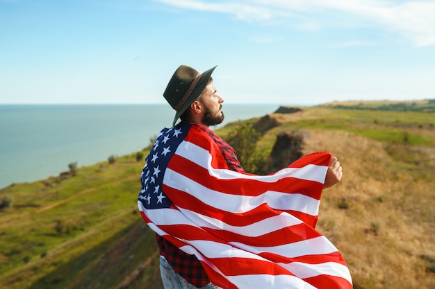 Foto 4 juli. vier juli. amerikaan met de nationale vlag. amerikaanse vlag. onafhankelijkheidsdag. patriottische vakantie. de man draagt een hoed, een rugzak, een overhemd en een spijkerbroek.