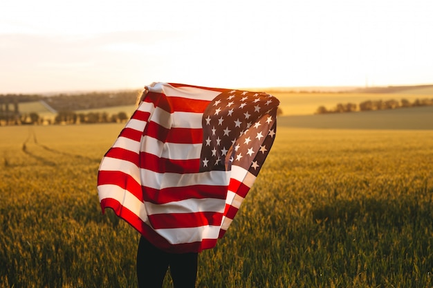 4 juli. Vrouw met de Amerikaanse vlag die in een tarwegebied bij zonsondergang loopt. Independence Day, patriottische vakantie.