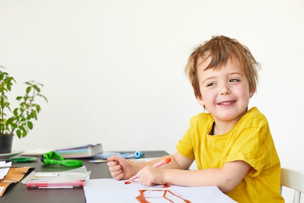 4-летний ребенок Милый мальчик рисует за столом и смотрит в камеру на белом