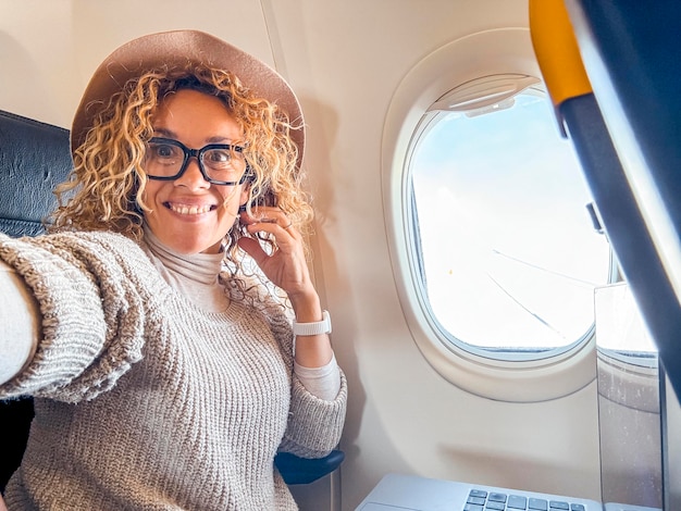 Foto 45-jarige blonde vrouw met een bril zit op een stoel in het vliegtuig, glimlacht en neemt een selfie en geniet van de tijd tijdens een vakantie reis reisconcept