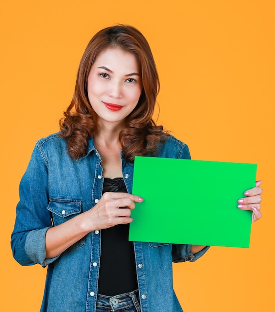 45s schattig mooi krullend haar Aziatische vrouw met groen blanco papier karton, studio-opname met flitslicht op felgele achtergrond. Idee voor advertentie-inhoud toegevoegd.