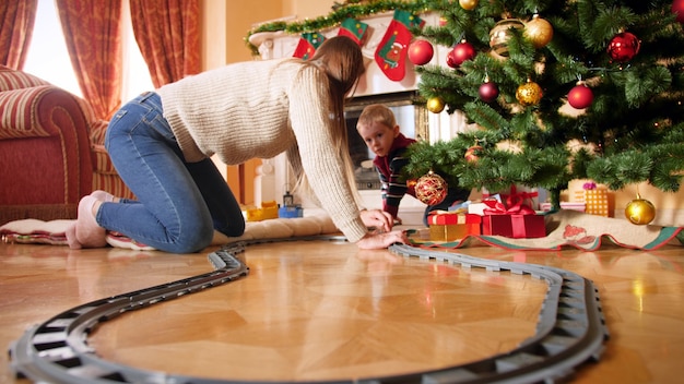 4k-beelden van een gelukkig lachend gezin met een kleine jongen die een spoorweg bouwt voor een kleine speelgoedtrein rond de kerstboom. Kind krijgt cadeaus en cadeautjes van de kerstman tijdens wintervakanties en vieringen