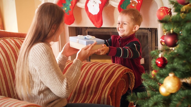 4k-beelden van lachende kleine jongen die kerstcadeau geeft aan zijn moeder. familie die nieuwjaarscadeautjes geeft en ontvangt in de woonkamer naast de kerstboom.