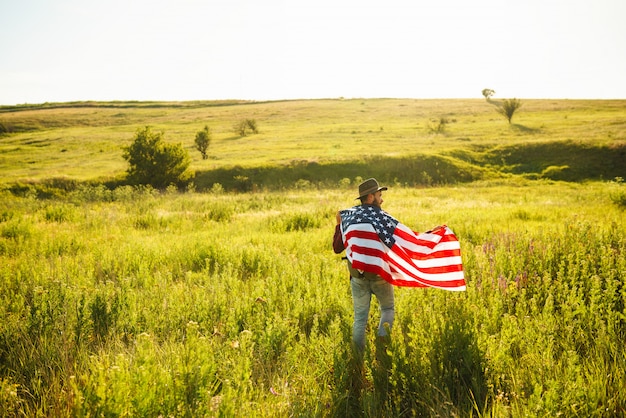 4th of July. Fourth of July. American with the national flag. American Flag. Independence Day. Patriotic holiday. The man is wearing a hat, a backpack, a shirt and jeans. 