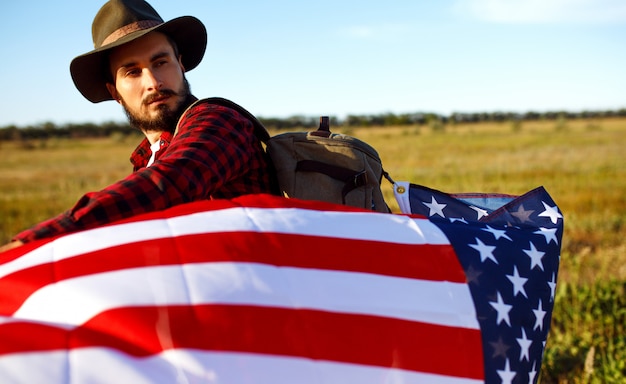 7月4日。 7月4日。国旗を持つアメリカ人。アメリカの国旗。独立記念日。愛国的な休日。男は帽子、バックパック、シャツ、ジーンズを着ています。