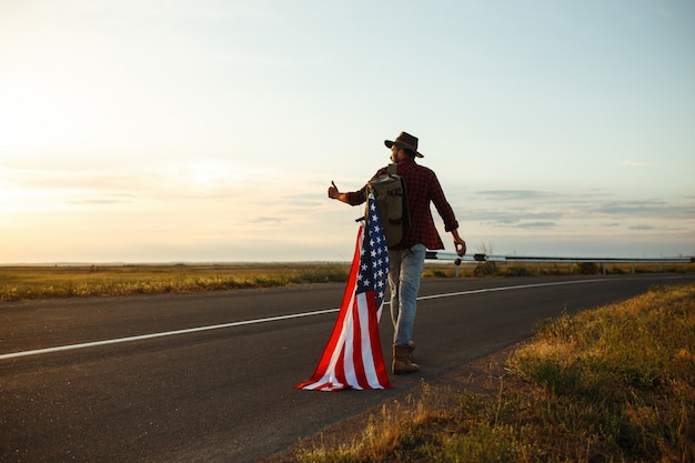 4 июля. Четвертое июля. Американец с национальным флагом. Американский флаг. День независимости. Патриотический праздник. Мужчина одет в шляпу, рюкзак, рубашку и джинсы.