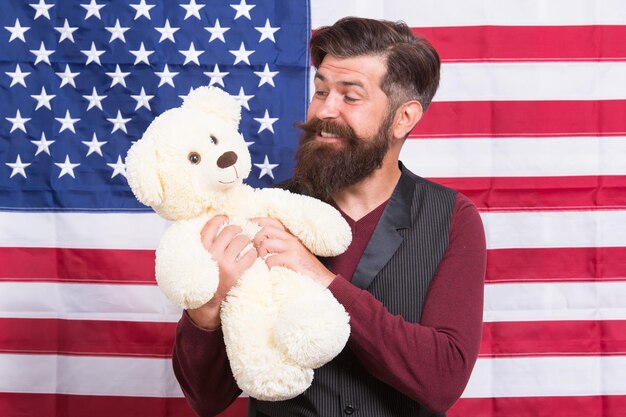 4th of July Independence day American guy with toy celebrate holiday American holiday Man bearded hipster hold teddy bear american flag background Child care Still childish Infant and funny