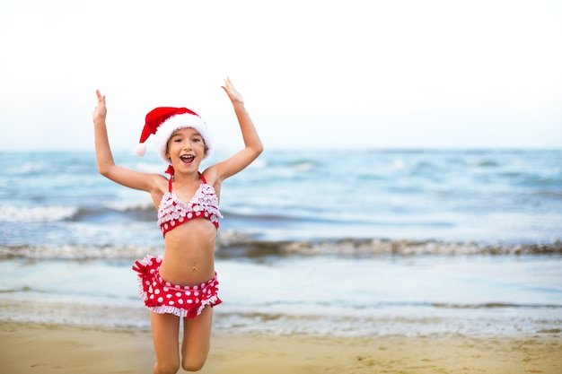 5-jarig meisje in rode zwembroek en kerstmuts op het strand is blij, schreeuwt, springt en zwaait met de handen van geluk. Kerst- en nieuwjaarswintertochten naar warme landen. Toerisme voor vakanties in de tropen