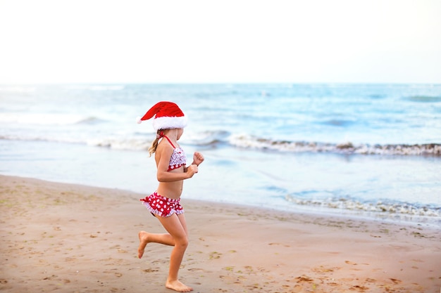 5-year-old girl in red swimsuit and Santa hat on beach is happy, screams, jumps and waves hands with happiness. Christmas and New Year winter tours to warm countries. Tourism for holidays in tropics