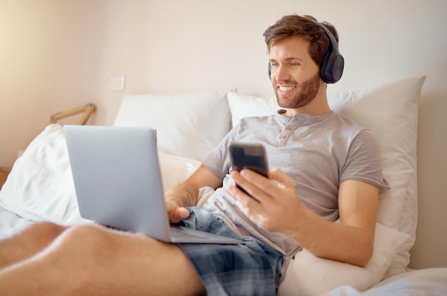 Video internet 5g e streaming online di un uomo su un computer e telefono a letto ragazzo felice con un sorriso rilassati e multitasking ascoltando un podcast radiofonico digitale o musica web con la tecnologia moderna