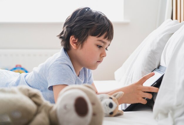 6-7-jarige jongen liggend met pluizig speelgoed en cartoon kijken op tablet, Gelukkig kind liggend in bed spelend spel in de ochtend, Kind met plezier en ontspannen in zijn eentje in de slaapkamer,