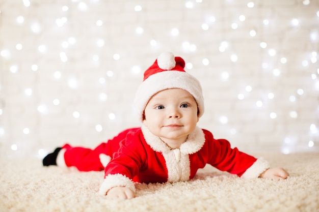 6 month old baby in Santa Claus costume is lying and smile over defocused lights