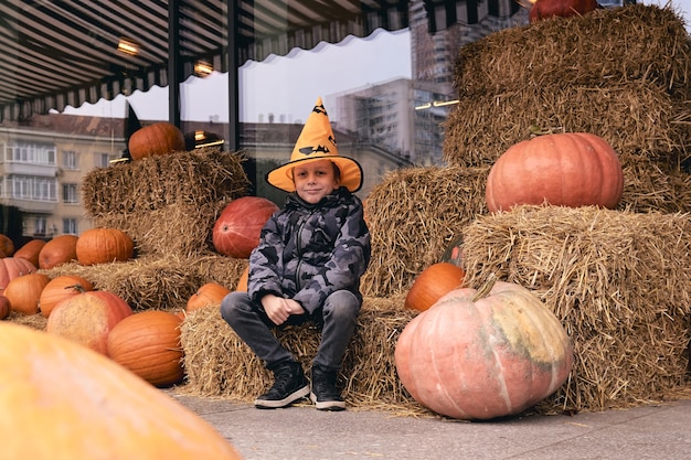 6 years boy in Halloween costume with pumpkins at farm market stands on sheaves of hay. Scary decorations. Kids trick or treat.