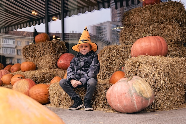 ファーマーズマーケットでカボチャとハロウィーンの衣装を着た6歳の少年は、干し草の束の上に立っています。怖い装飾。子供たちはトリックオアトリート。