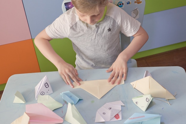 6歳の少年は、検疫Covid-19、自己隔離、オンライン教育の概念、ホームスクーリング中に折り紙の飛行機とカエルを作ります。家にいる子供、幼稚園は閉鎖、キッズアート。