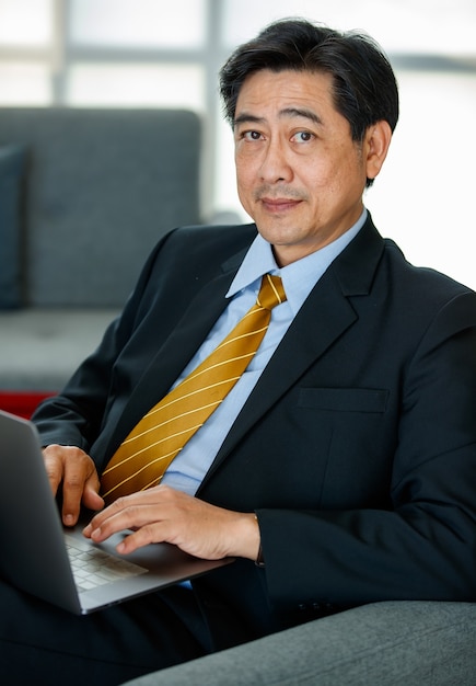 Photo 60s asian senior successful executive business male entrepreneur sitting, leaning back on sofa in the indoor office, using laptop for technology, working with confidence, smiling with positivity.