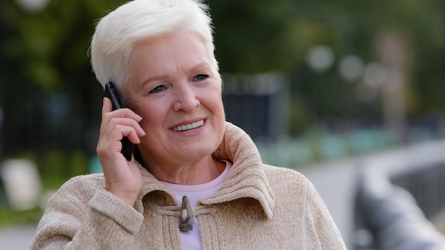 60s blonde woman holds phone chatting with friend outdoors having pleasant distant conversation talking with grown up children elder generation and modern technologies everyday easy usage concept