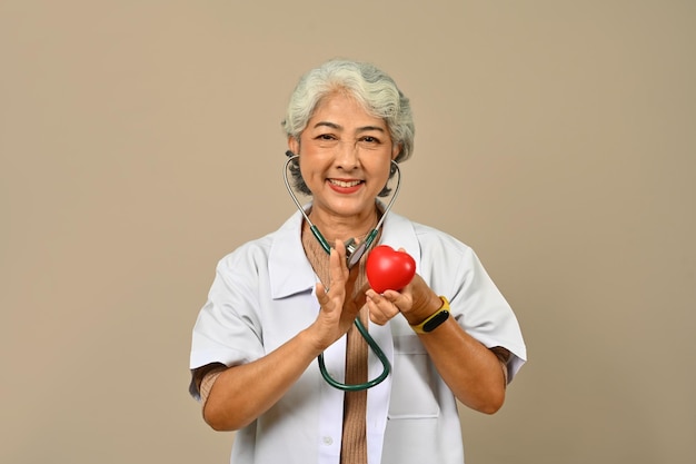 60s mature female doctor holding red heart over beige background Cardiology medicine donation and healthcare concept