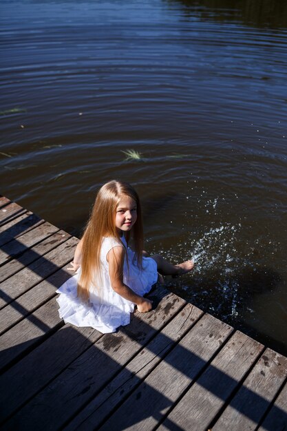 湖のほとりに長いブロンドの髪をした7歳の少女が、足を水につけてクラッチに座っています。彼女は湖に足をはねかける。長い髪の白いドレスを着た裸足の女の子。