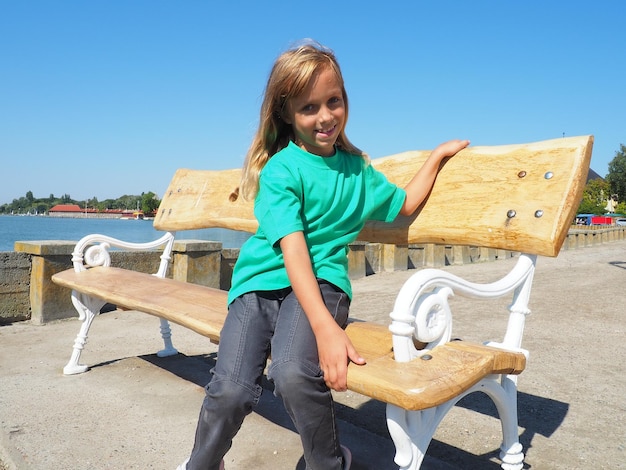 A 7 years old white caucasian girl in a green Tshirt sits on a wooden bench embankment of Lake Palic Serbia A child smiles on a bench in sunny summer weather Tourism and children's vacations