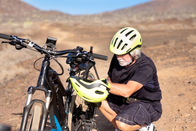 70 year old man with yellow helmet checks his electric bike Outdoor activity in arid country road