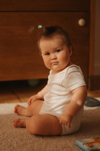 A 7month girl is sitting near a dresser in a bodysuit at home