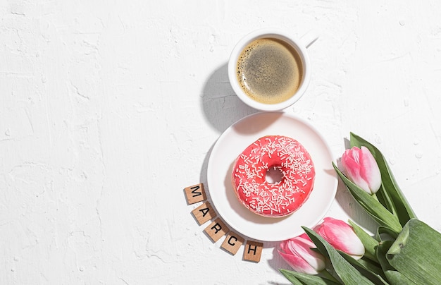 Biglietto di auguri dell'8 marzo con una tazza di caffè e un piattino con una ciambella che forma il numero otto e un tenero tulipano rosa nelle vicinanze