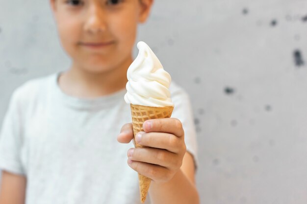 8 year old boy holds out vanilla ice cream in waffle cone on grey background, selective focus