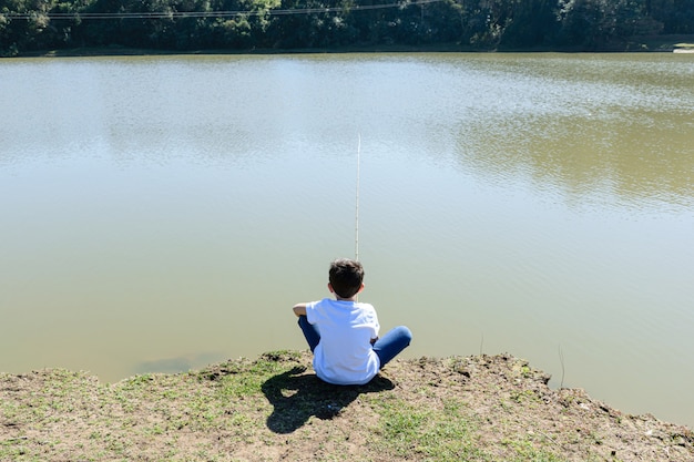 晴れた朝、湖のほとりで釣りをしている、背を向けて座っている8歳のブラジル人の子供。