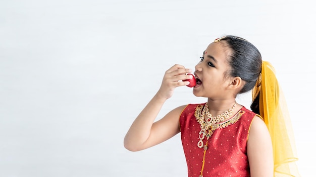 8 year old Indian girl wearing a red sari holding and eating fruit is an apple