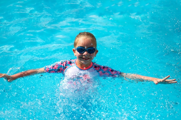 8-летняя девочка в ярком купальном костюме и синих очках стоит в бассейне под солнцем с голубой водой