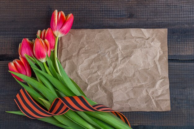 9 May background - template blank greeting card with red tulips, George ribbon and paper note on the wooden background. Victory day or fatherland defender day concept. Top view, copy space for text