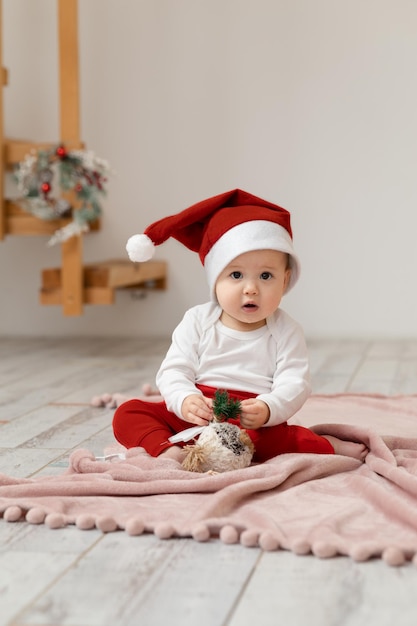크리스마스 의상을 입은 9개월 된 아시아 아기가 바닥에 깔린 담요 위에 새해 장난감을 가지고 놀고 눈을 크게 뜨고 카메라를 쳐다봅니다